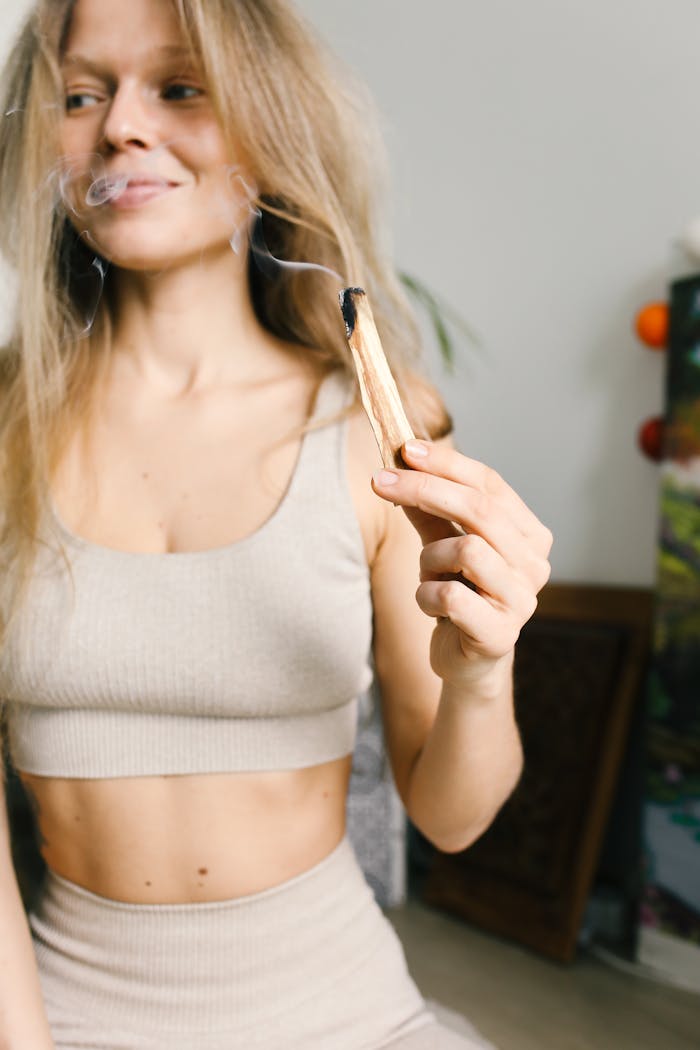 Young woman smiling while holding a burning sage stick indoors, implying meditation and wellness.